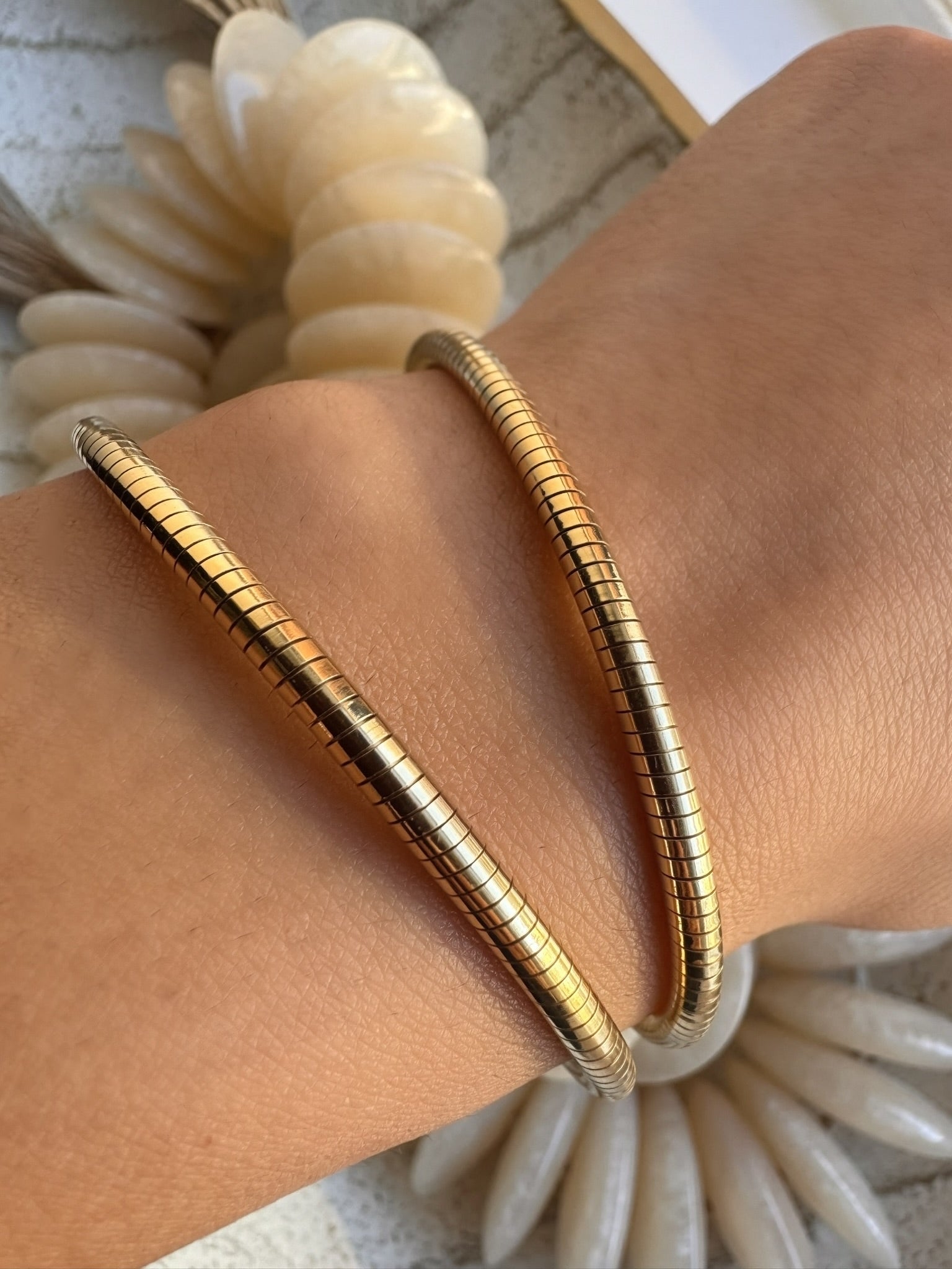 Two slim gold snake-bangle bracelets worn on a person's wrist, shown close-up against a light neutral background with decorative shell-like elements.